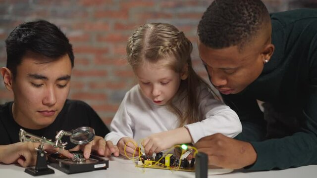 Schoolgirl and black man engineer work with chip studying electricity effect and green lamp lights while Asian guy fixes hard disk drive at table closeup