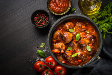Meatballs in tomato sauce in black skillet on dark stone table with ingredients. Top view copy space.