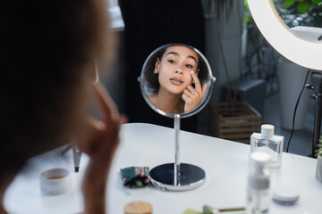 Blurred african american woman applying cosmetic cream near mirror and perfume at home.