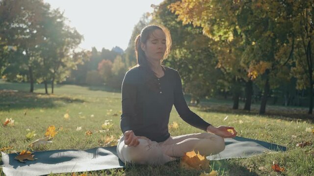Young woman sitting on mat in lotus position in city park on autumn day. Sunny autumn day in a city park. Sporty girl practicing meditation outdoors. High quality 4k footage