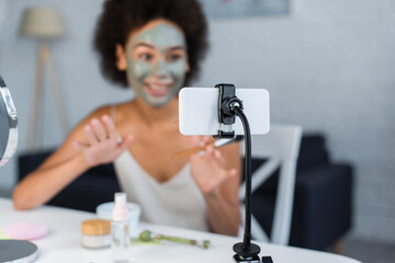 Smartphone near blurred african american blogger with clay mask on face at home.
