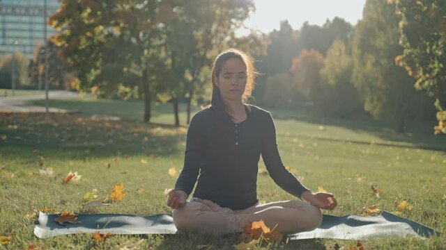 Young woman sitting on mat in lotus position in city park on autumn day. Sunny autumn day in a city park. Sporty girl practicing meditation outdoors. High quality 4k footage