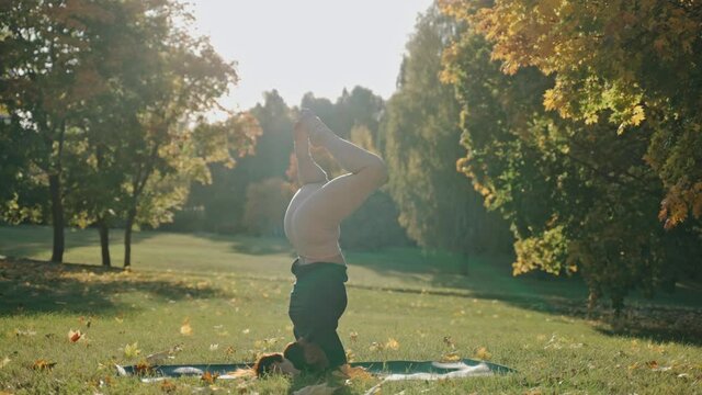 Young woman practicing headstand yoga pose in city park on sunny autumn day. Sunny autumn day in a city park. Sporty girl practicing meditation outdoors. High quality 4k footage
