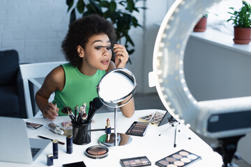 Pretty african american blogger applying mascara near mirror, gadgets and decorative cosmetics at home.