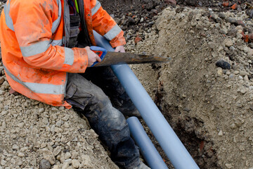 Groundworker in hi-viz coat cutting grey plastic telecom duct with hand saw while sitting at the edge of trench