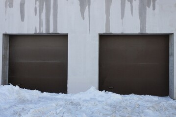 gray concrete wall of a garage with wet spots and two brown metal gates on a winter street in drifts of white snow