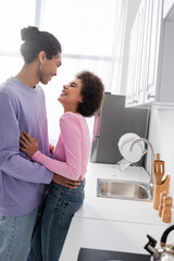 Side view of positive african american woman hugging boyfriend near worktop in kitchen.