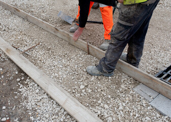 Groundworker in orange safety hi vis trousers fixing a timber along string line with steel pin to form a kerb riser and a straight edge for tarmac road surface during new road construction