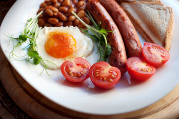 English breakfast with scrambled eggs, sausages, toast, tomatoes, microgreens and beans on a white plate. Breakfast on a wooden board