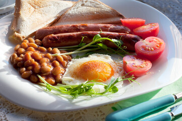 English breakfast with scrambled egg, sausages, toast, tomatoes, pea sprouts and beans on white plate. Traditional English breakfast  with knife and fork on white lace tablecloth