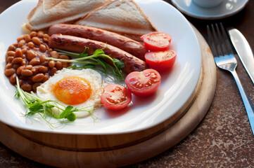 English breakfast with scrambled eggs, sausages, toast, tomatoes, pea sprouts and beans on a white plate. Traditional english breakfast with tea, fork and knife on a dark brown background