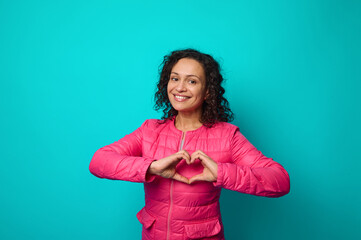 Emotional portrait of beautiful woman in pink jacket smiling toothy smile looking at camera, holding hands on her chest and showing heart shape with her hand, isolated on blue background, copy space