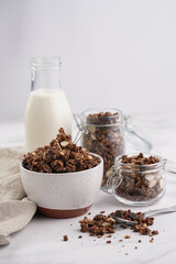 Breakfast muesli cereal with chocolate chips and nuts in several jars and bowl, bottle of milk, white surface