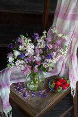 Background with a bouquet in a spring and summer vase of pink, white, purple flowers, aquilegia, mattiola and a plate with strawberries, a shawl on a chair, against the wooden background, still life.