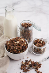 Breakfast muesli cereal with chocolate chips and nuts in several jars and bowl, bottle of milk, white surface