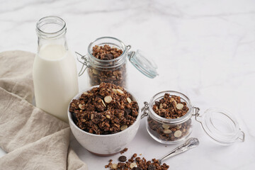 Breakfast muesli cereal with chocolate chips and nuts in several jars and bowl, bottle of milk, white surface