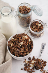 Breakfast muesli cereal with chocolate chips and nuts in several jars and bowl, bottle of milk, white surface