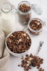 Breakfast muesli cereal with chocolate chips and nuts in several jars and bowl, bottle of milk, white surface
