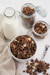 Breakfast muesli cereal with chocolate chips and nuts in several jars and bowl, bottle of milk, white surface