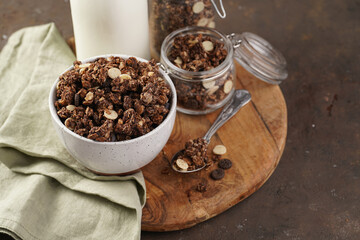 Breakfast muesli cereal with chocolate chips and nuts in several jars and bowl, bottle of milk, brown wooden surface