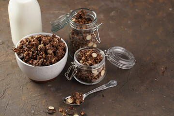 Breakfast muesli cereal with chocolate chips and nuts in several jars and bowl, bottle of milk, brown wooden surface