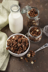 Breakfast muesli cereal with chocolate chips and nuts in several jars and bowl, bottle of milk, brown wooden surface