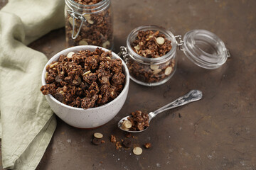 Breakfast muesli cereal with chocolate chips and nuts in several jars and bowl, bottle of milk, brown wooden surface