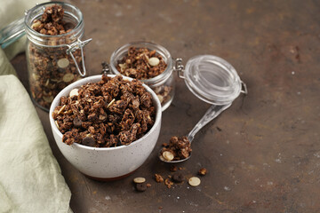 Breakfast muesli cereal with chocolate chips and nuts in several jars and bowl, bottle of milk, brown wooden surface