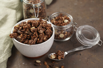 Breakfast muesli cereal with chocolate chips and nuts in several jars and bowl, bottle of milk, brown wooden surface