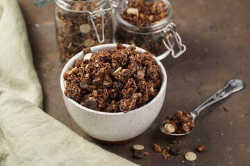 Breakfast muesli cereal with chocolate chips and nuts in several jars and bowl, bottle of milk, brown wooden surface
