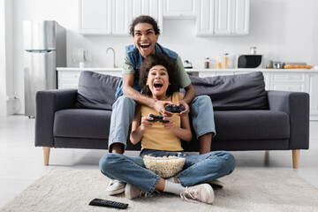 Obraz premium KYIV, UKRAINE - NOVEMBER 9, 2021: Excited african american couple with popcorn playing video game at home.