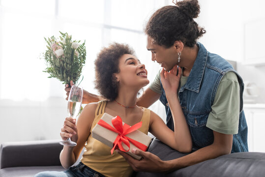 Positive woman holding champagne near african american boyfriend with present and bouquet at home.