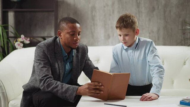 Skilled African-American teacher shows exercise in textbook to junior student at private English language tuition with native speaker slow motion