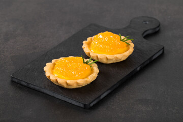 Tartlets with custard and slices of tangerines covered with syrup on a serving board on a dark gray background
