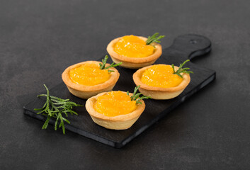 Dessert small Tartlets with custard and slices of tangerines covered with syrup on a serving board on a dark gray background