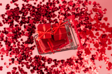 top view of small shopping cart with presents near shiny red confetti hearts on pink.