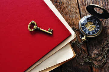 A top view image of a single vintage key on a bright red book along with an antique pocket watch. 