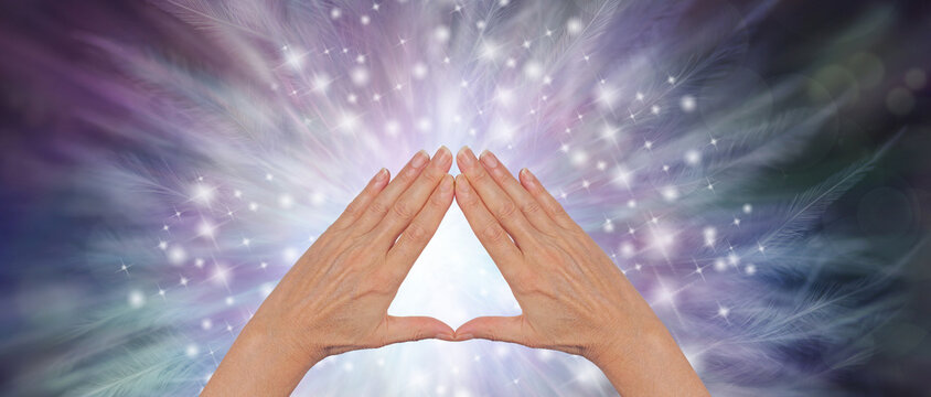 The triangle is a stunning healing tool  - female hands making a triangle against a wide sparkling feathered ethereal background with white light inside the triangle and copy space both sides
