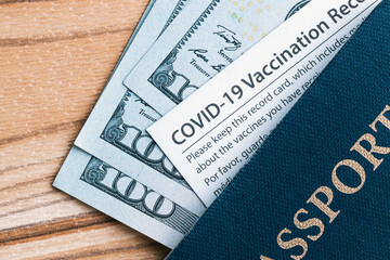 One Covid-19 Coronavirus vaccination card inside a blue American passport with some one hundred dollar bills cash requirement mandate law