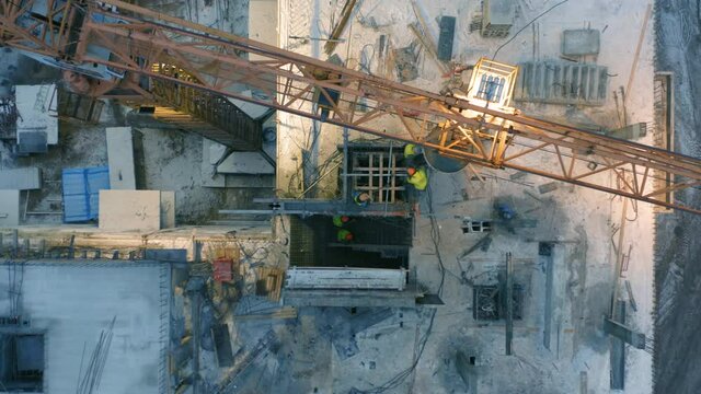 Aerial top down view of a tower crane with a concrete bucket is unloaded onto a rooftop. Workers control the concrete pouring on a floor of an apartment building under construction at winter dusk
