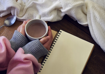 A hand holding a cup of hot chocolate, beside it is a blank page of a note book for copy space.