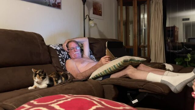 Shirtless man sits on couch, looks at camera and smiles. Pets cat.