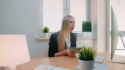 Elegant blond female sitting at desk at workplace with computer holding in hands bundle of cash looking astonished at monitor, and throwing money in air and happily making yes gesture.