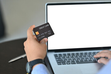 Cropped image of a young businessman holding a credit card for doing an online payment in front of a computer laptop at the working desk.