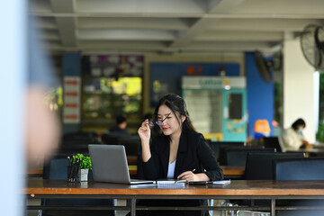 Photo of a young businesswoman wearing eyeglasses while sitting at the wooden working desk surrounded by a computer laptop and documents.