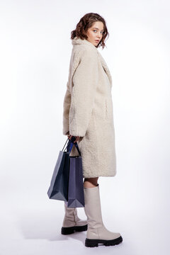 High fashion photo of a beautiful elegant young woman in a pretty cream beige fur coat, boots, two blue shopping bags posing over white background. Make up, hairstyle. Slim figure.