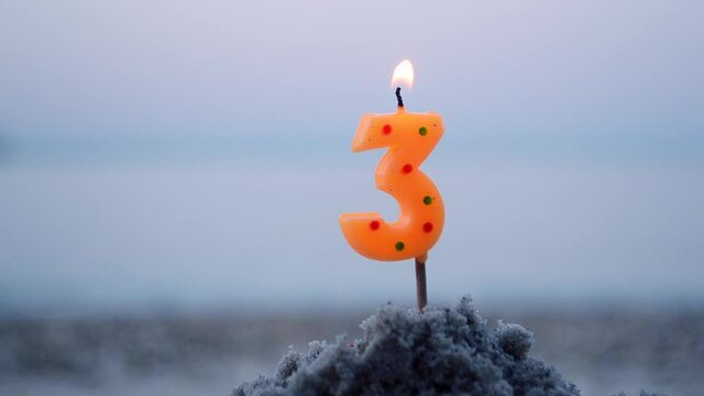 Candle number three on stick inserted into sand and burns. Lighted candle in sand on background of blurry sea waves. Green burning candle on blurred blue brown background. Birthday Celebration Concept