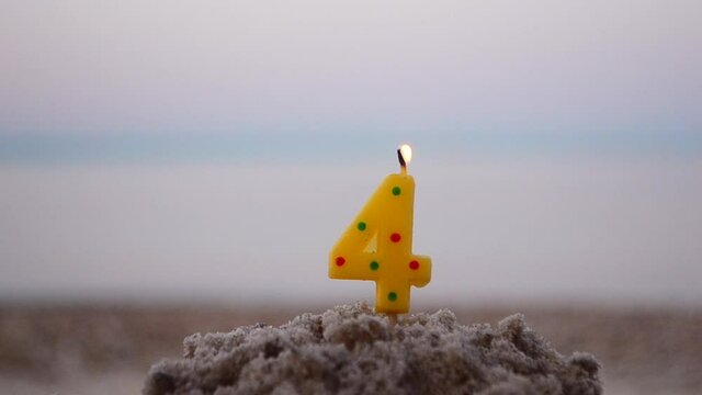 Candle number four on stick inserted into sand and burns. Lighted candle in sand on background of blurry sea waves. Green burning candle on blurred blue brown background. Birthday Celebration Concept