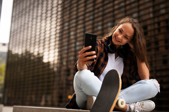 Fashion woman with skateboard. Young happy girl using the phone. Beautiful woman enjoy outdoors