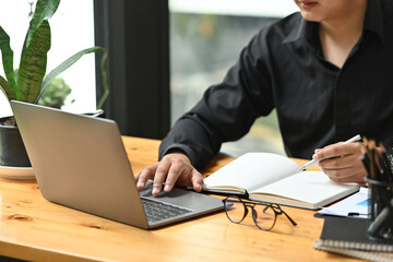 Cropped image of a young office man writing on a notebook while using a computer laptop at the wooden working desk.